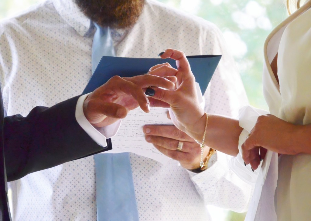 Close-up of a bride's hand with dark manicured nails placing a wedding ring on the groom's finger while an officiant holds a blue folder containing written vows in the background.
