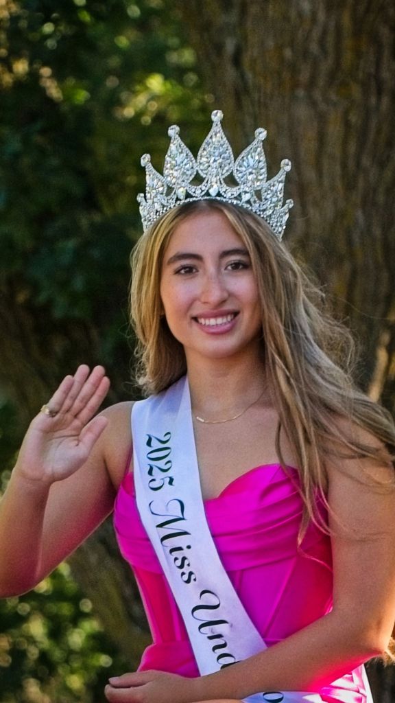 A close-up portrait of the 2025 Miss Underwood title holder smiling and waving while seated during the 2026 Underwood Harvest Festival Parade. She wears a brilliant, jewel-encrusted silver tiara, a bright pink gown, and a white sash reading "2025 Miss Underwood" as she raises her right hand in a wave against a soft-focus background of green trees.