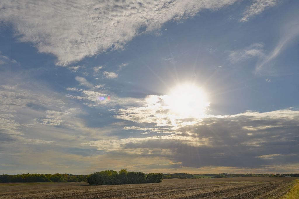 A dramatic wide-angle landscape photograph of a brilliant sun bursting through dynamic clouds over a tilled agricultural field in rural Minnesota, with bright sun rays casting across an expansive blue and white sky.