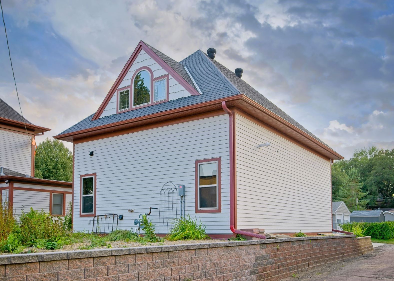Three-quarter corner view of the east and rear facades of the matching carriage house garage at 504 W. Cavour Ave in Fergus Falls, Minnesota, showcasing red trim lines and a retaining wall.