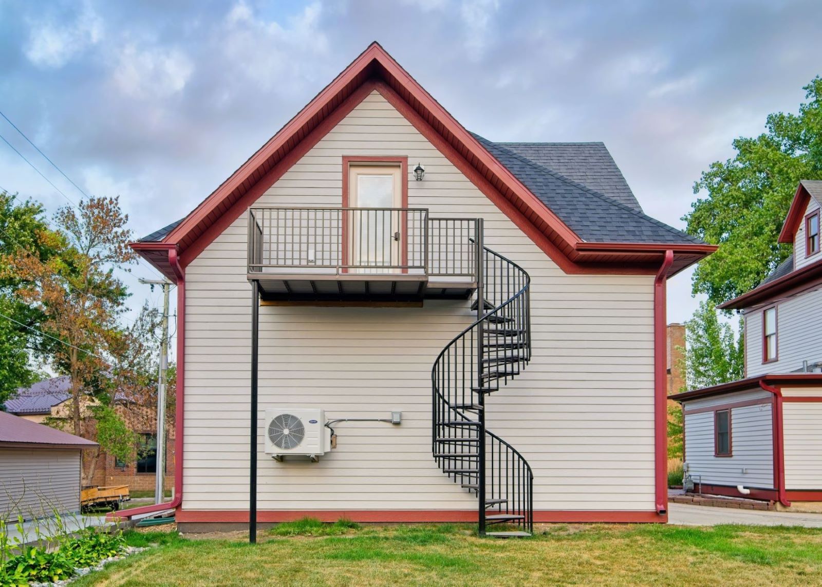 West side exterior view of the detached carriage house at 504 W. Cavour Ave in Fergus Falls, Minnesota, showcasing a black spiral staircase, second-story balcony, and matching red trim lines.