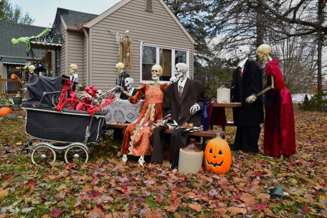 An elaborate outdoor Halloween display featuring several life-sized skeletons dressed in formal vintage clothing gathered around a picnic table in a leaf-covered yard.