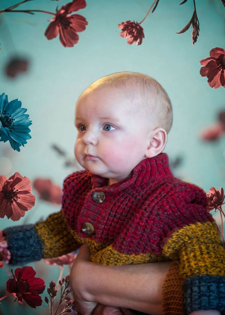 A wide-eyed baby wearing a colorful knit sweater looks off camera against a teal backdrop framed by floating maroon and blue flowers.