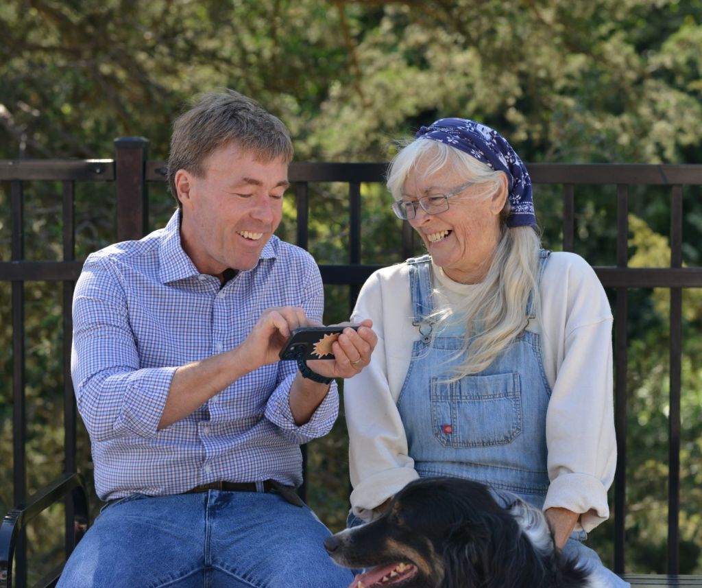 A candid, sunny outdoor photograph of Ben Schierer and Valerie Thompson sitting together on a bench, laughing as they look at a smartphone, with a black and tan dog in the foreground.