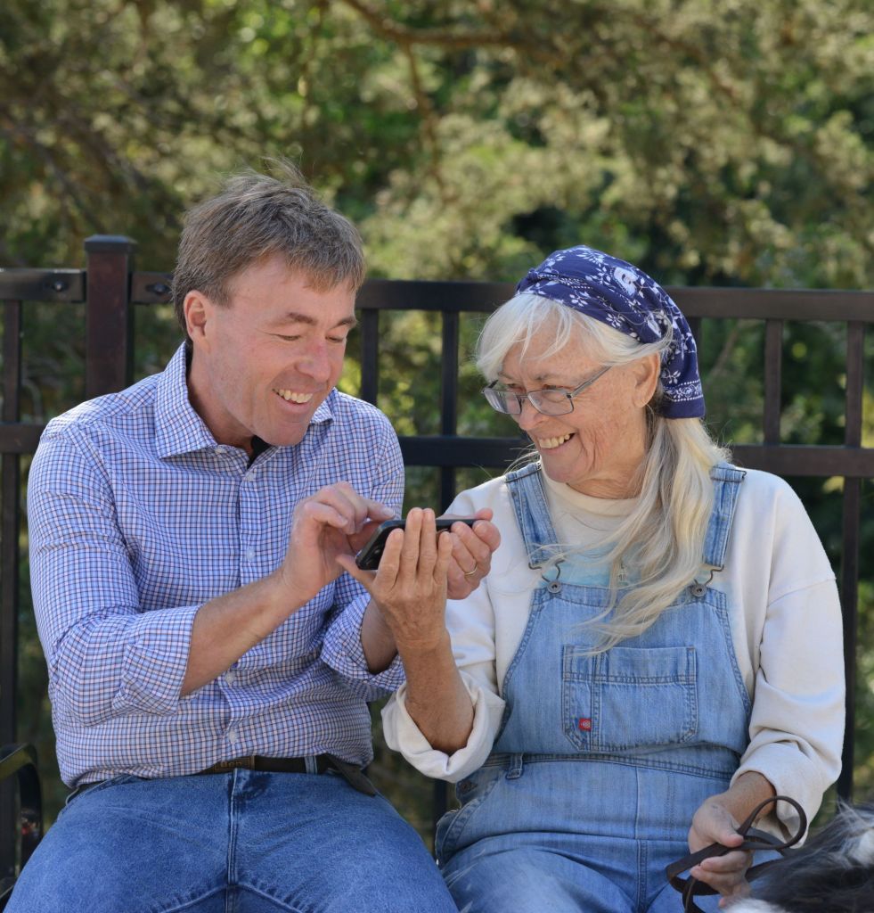 A close-up candid photograph of Ben Schierer and Valerie Thompson looking closely at a smartphone screen, both smiling warmly as Valerie holds the edge of the phone.