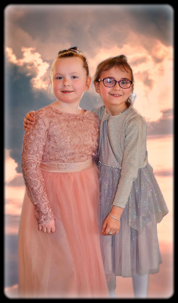 Two young girls standing arm-in-arm in formal party dresses smiling against a dramatic sunset background with pink and orange clouds.