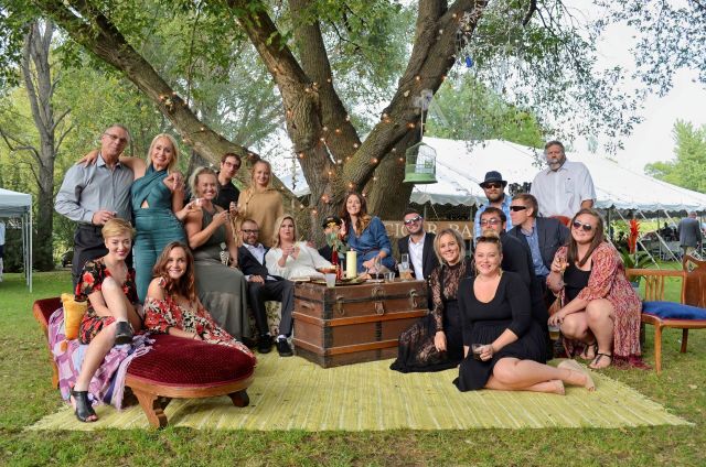 A large, stylishly dressed group of friends smile and pose together with the bride and groom at an upscale outdoor lawn party beneath a large tree. The Beck Wedding group portrait on a lawn under a lit tree, featuring the late Brian Beck, his daughter Samantha the bride, and wedding guests.