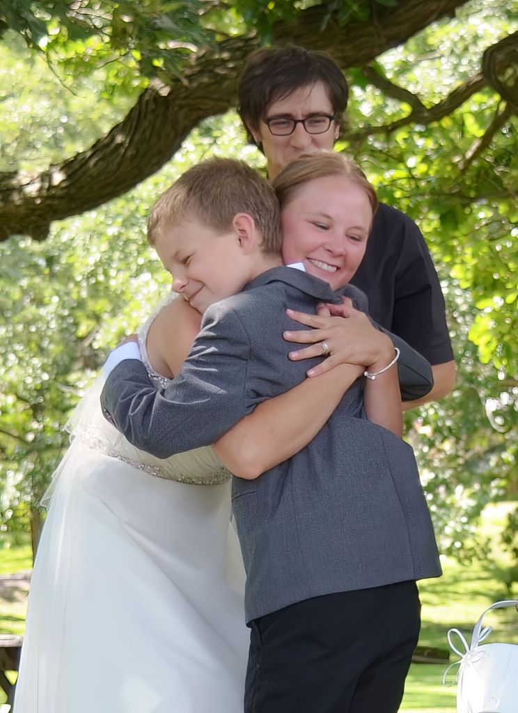 A smiling bride in a white wedding gown shares a warm hug with a young boy in a grey suit jacket during an outdoor ceremony while an officiant looks on.
