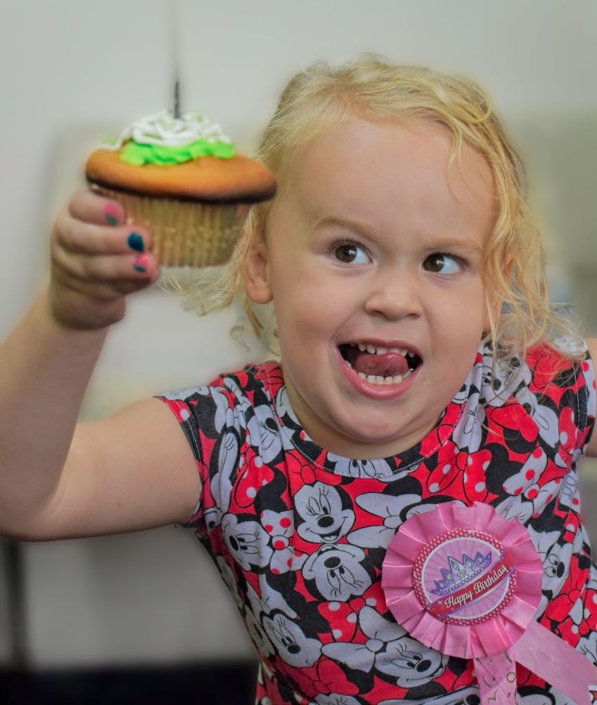 An excited young girl wearing a Minnie Mouse shirt and a pink birthday ribbon holds out a cupcake with green frosting.