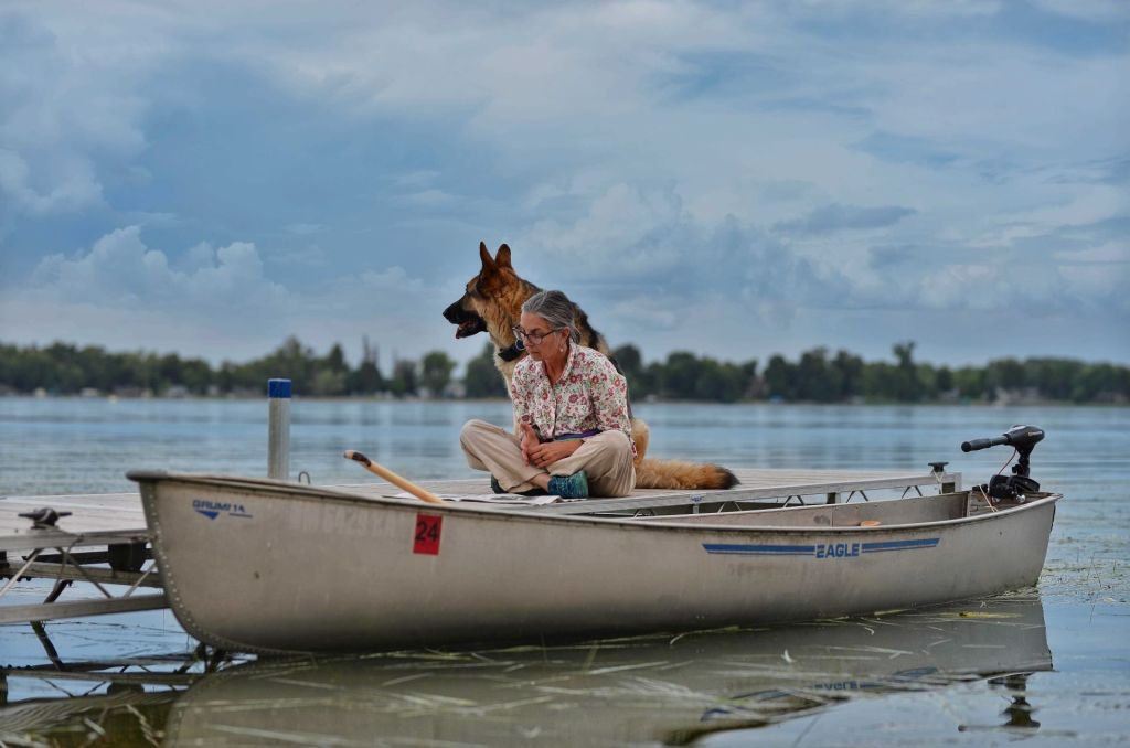 Bonnie Bell Albers sits cross-legged on a wooden dock next to a silver Grumman canoe with an electric motor, accompanied by a large German Shepherd dog at Peterson Point on Star Lake, MN.
