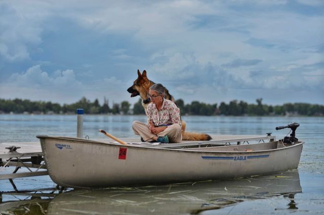 Bonnie Bell Albers sits cross-legged on a wooden dock next to a silver Grumman canoe with an electric motor, accompanied by a large German Shepherd dog at Peterson Point on Star Lake, MN.