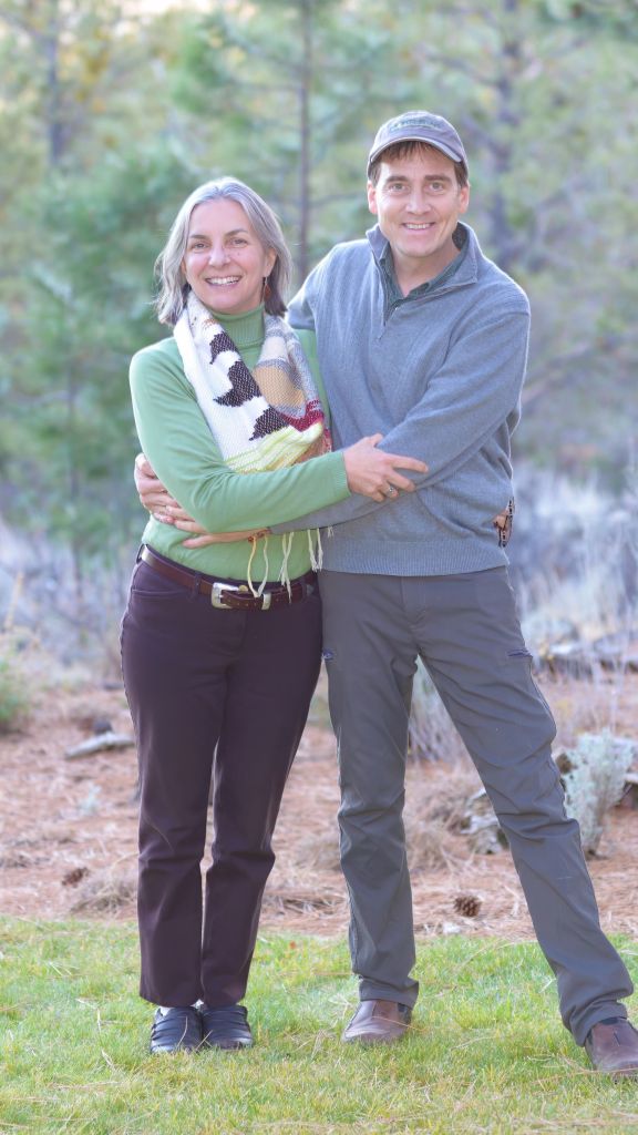 Fine art photographer Lance Albers and his wife Bonnie Bell Albers smiling together during an outdoor portrait session, standing on green grass with a soft pine forest background.