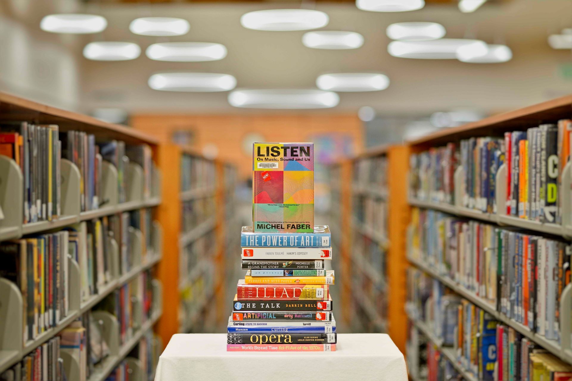 A stacked pillar of books centered between long rows of bookshelves inside the brightly lit Fergus Falls Public Library.