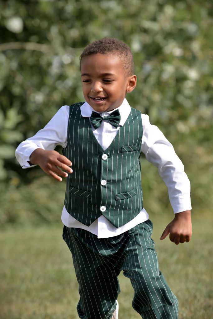 A young boy running outdoors wearing a green pinstripe vest, matching trousers, a white button-down shirt, and a dark green bow tie.