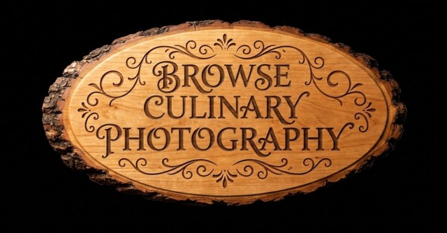 Oval dark walnut wood slice sign with bark edges engraved with the text "Browse Culinary Photography" in elegant serif script, framed by ornate flourish carvings on a solid black background.