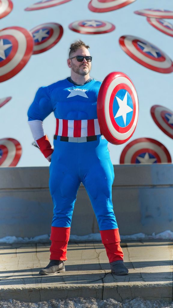 A man wearing sunglasses and a padded Captain America costume stands outside on a sunny day, holding a red, white, and blue shield on his left arm while dozens of digital Captain America shields float and blur in the background around him.