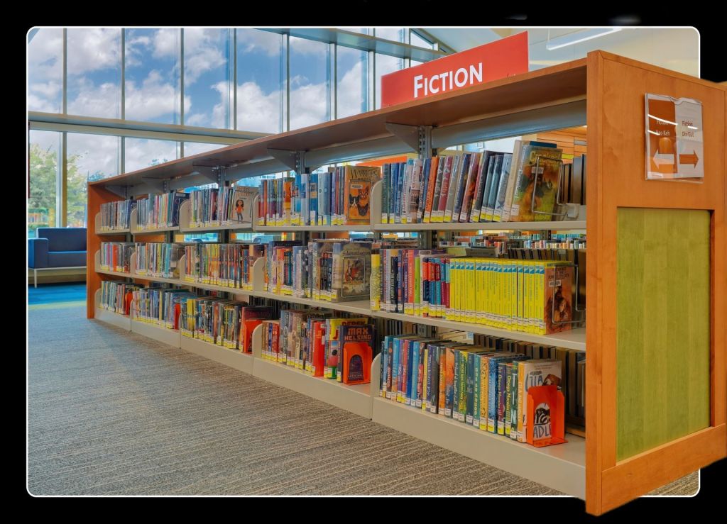 Brightly colored children's fiction books lined up on wooden bookshelves in front of a large window at the Fergus Falls Public Library.