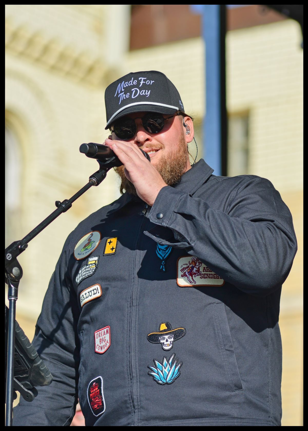 Musician Chris Tungseth smiling and singing into a microphone while wearing a patch-covered jacket and a black Made For The Day cap in Fergus Falls, Minnesota.