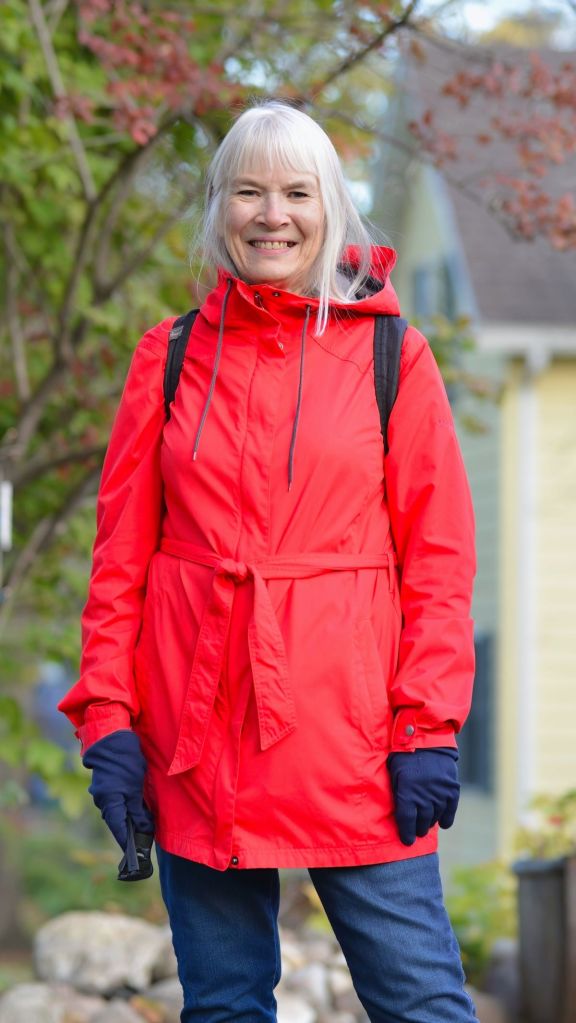 A smiling woman with long silver hair wearing a bright red hooded raincoat, dark blue gloves, and blue jeans outdoors.