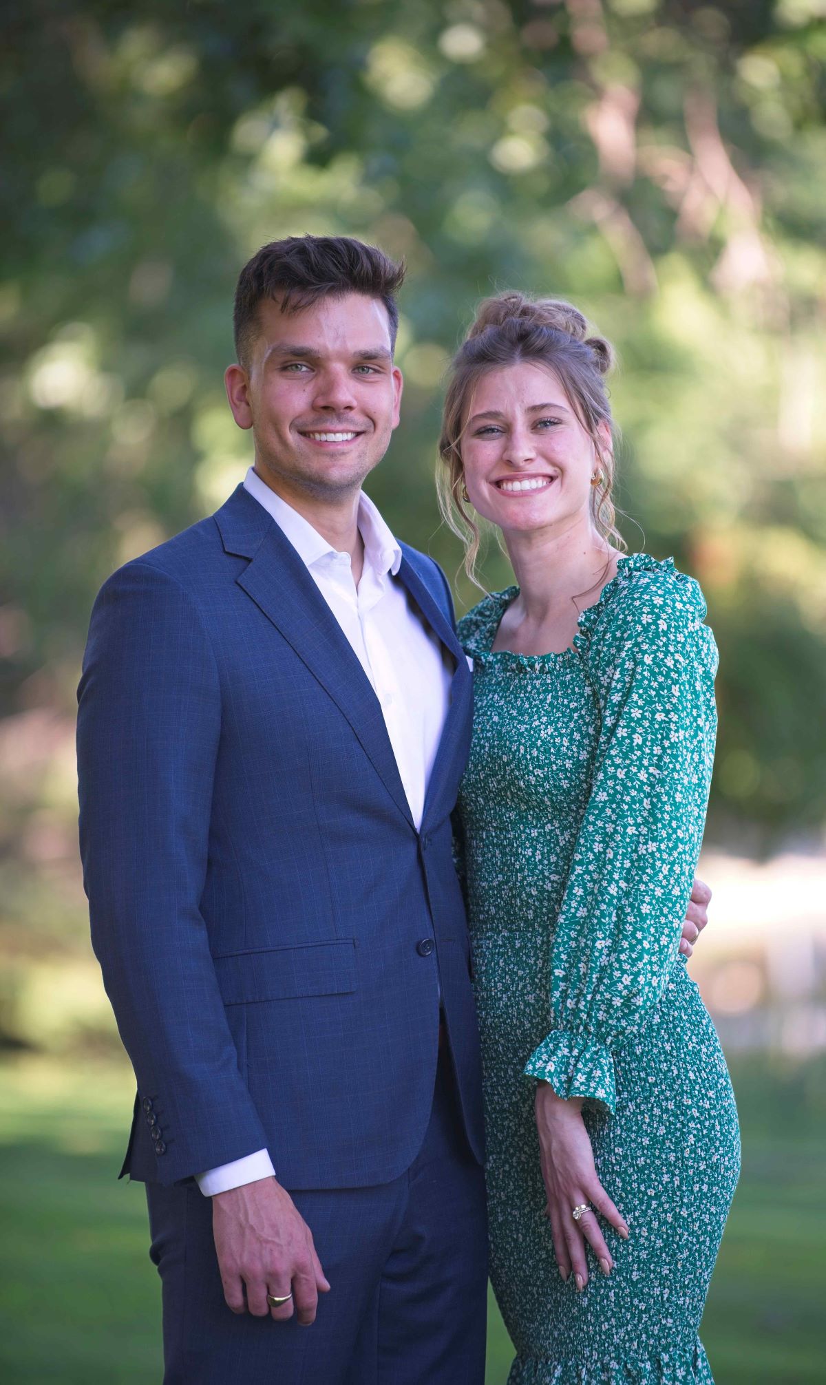 A smiling young couple stands close together outdoors; the man wears a blue suit jacket, and the woman wears a green floral dress.