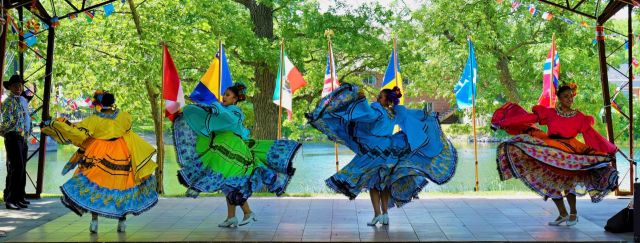 Four traditional Mexican folkloric dancers perform on an outdoor stage with large, vibrant skirts spread wide, framed by multi-national flags and a river in the background.