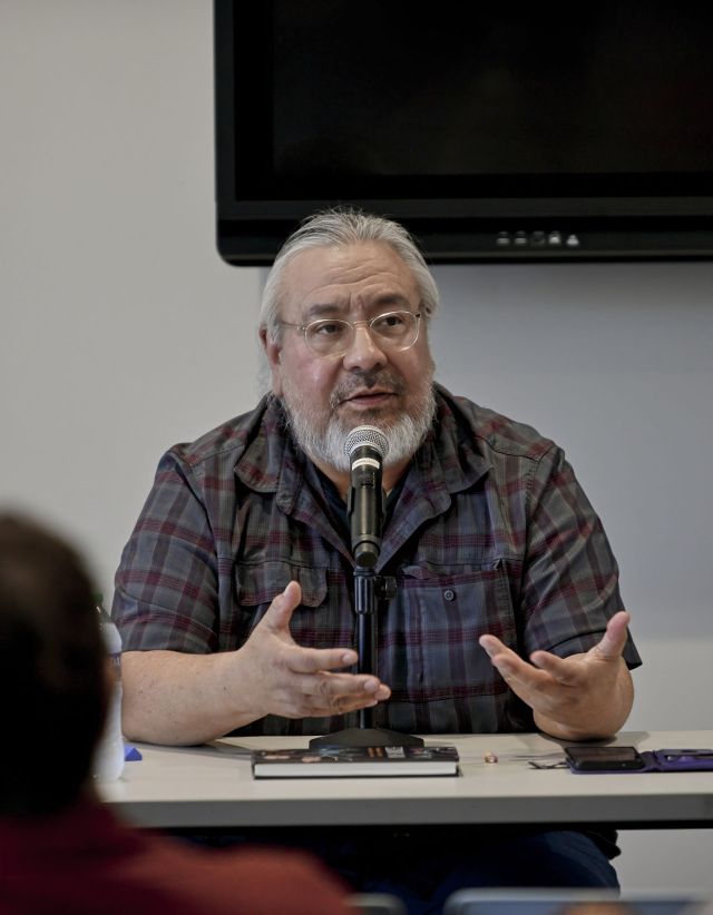 Darren Renville speaking into a microphone with expressive hand gestures at the Doctrine of Discovery presentation inside the Fergus Falls Public Library.