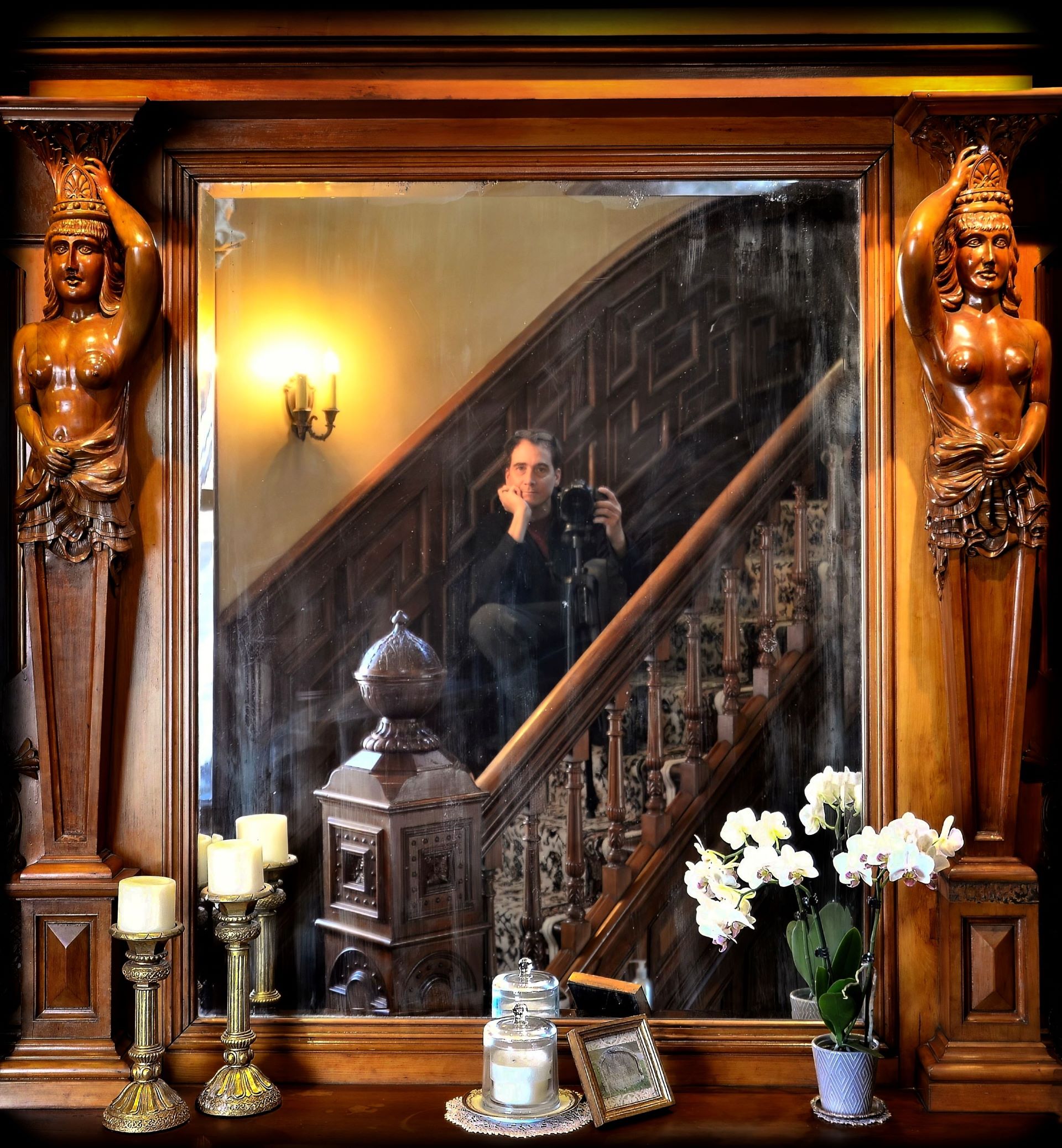 Photographer Lance Albers holding a camera, captured in a mirror reflection inside the historic Dearing Mansion in St. Paul, MN, framed by a carved wooden mantel and a grand staircase.