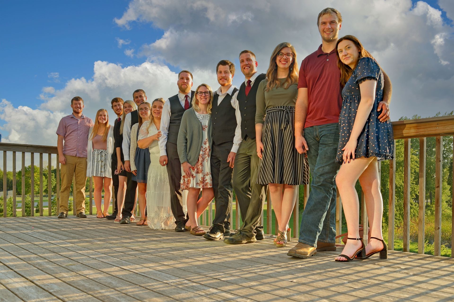 A large wedding party or family group standing in a diagonal line on an outdoor wooden deck at The Barn venue in Dunvilla, Minnesota, under a blue sky with white clouds.