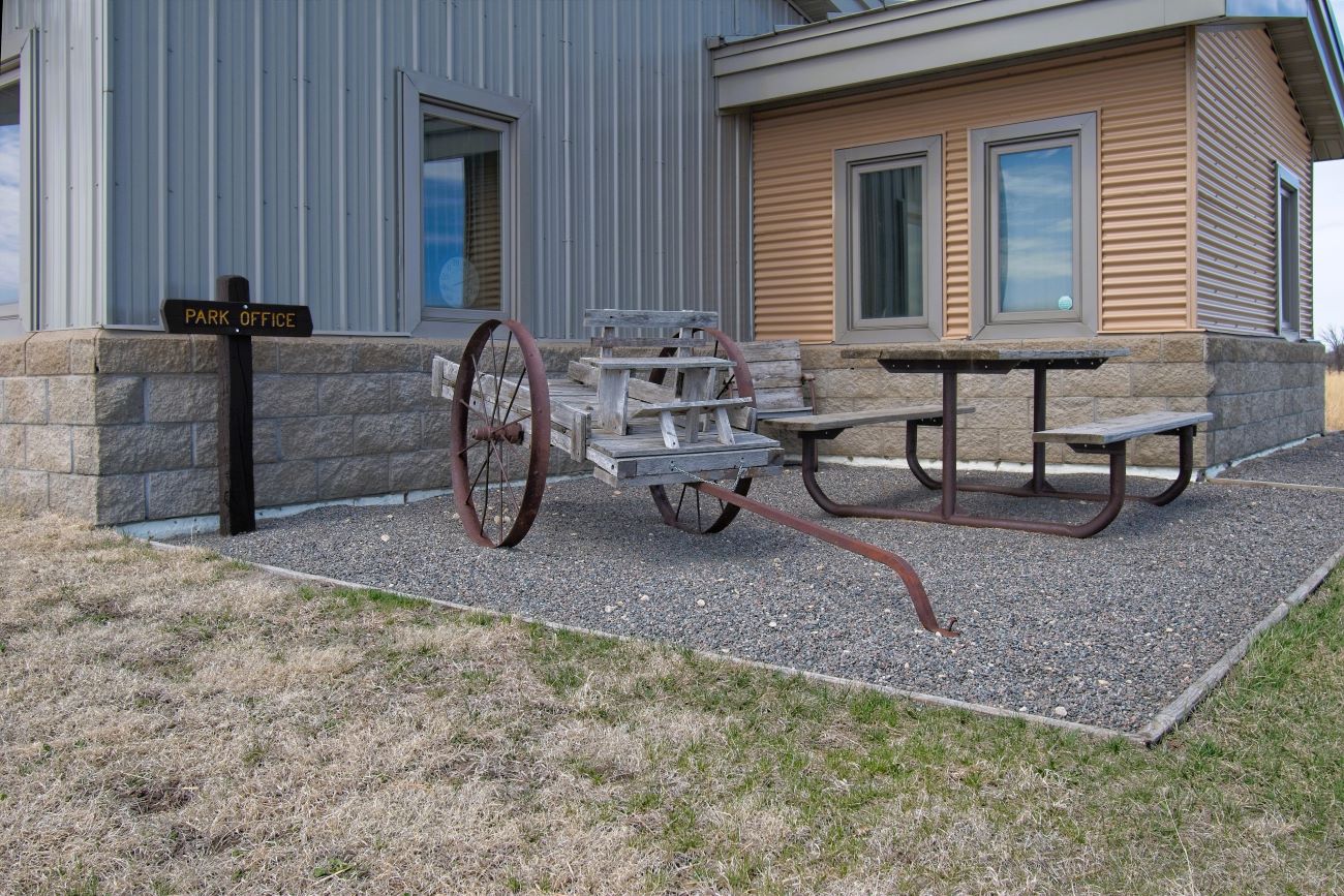 An antique wooden buckboard wagon display next to a wooden picnic table outside a park office building with gray and tan metal siding.