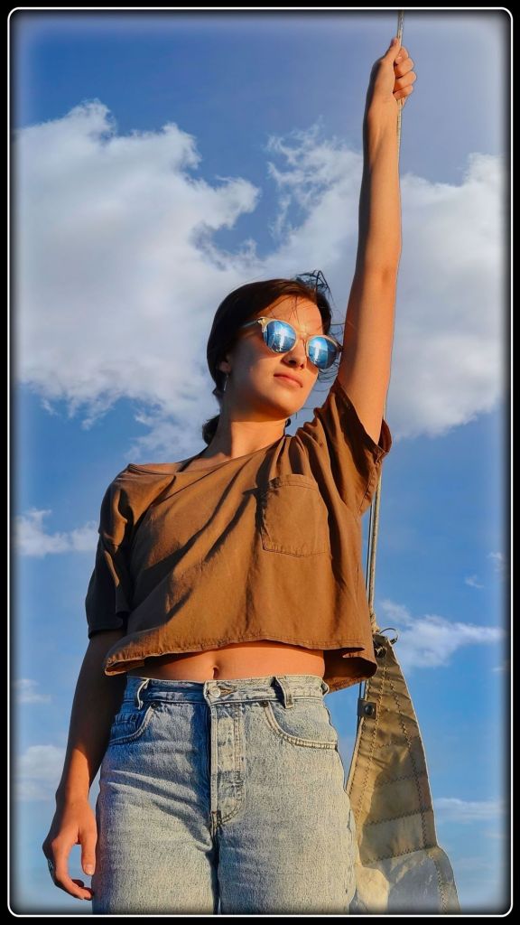 A young woman in a brown crop top, jeans, and mirrored sunglasses stands on a sailboat, holding a rope against a blue sky.
