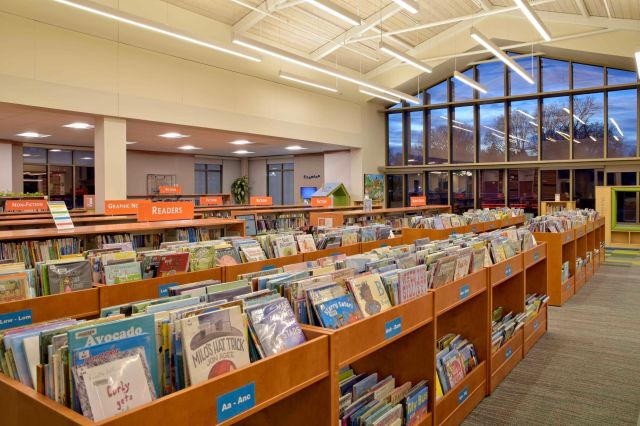 The children's section of the Fergus Falls Public Library featuring rows of wooden bookshelves filled with picture books beneath a high vaulted ceiling and large windows.