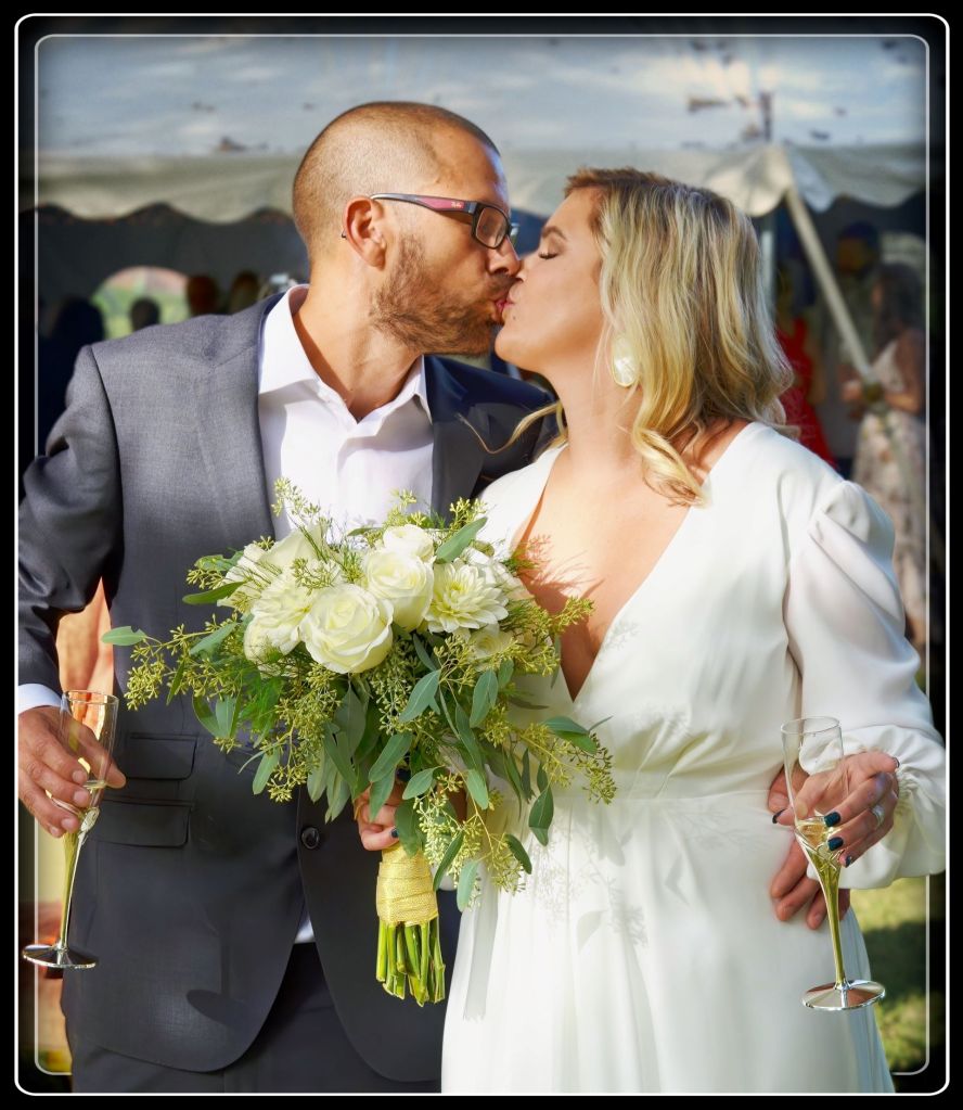 A bride and groom sharing a romantic kiss while holding champagne flutes and a lush white rose bouquet at a luxury tented wedding reception in Otter Tail County, Minnesota.