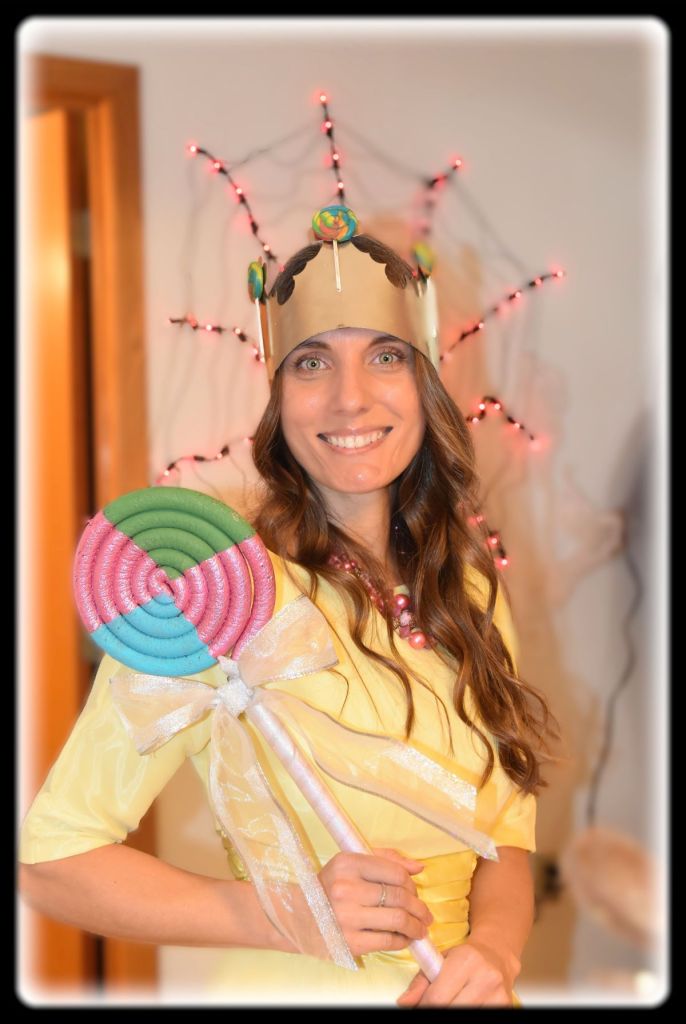 A smiling woman wearing a gold crown decorated with small lollipops, dressed in a yellow top, and holding a large, colorful prop lollipop with a white ribbon bow.