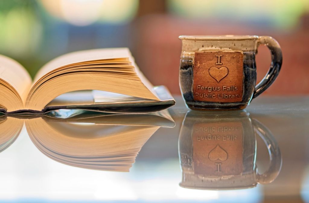 An open hardcover book and a handmade ceramic mug reading "I [Heart] Love Fergus Falls Public Library" reflecting on a glossy glass surface with a soft, blurred background.