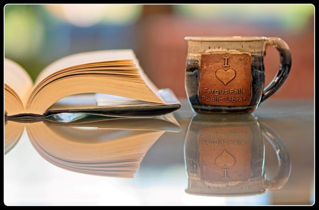 An open hardcover book and a handmade ceramic mug reading "I [Heart] Love Fergus Falls Public Library" reflecting on a glossy glass surface with a soft, blurred background.
