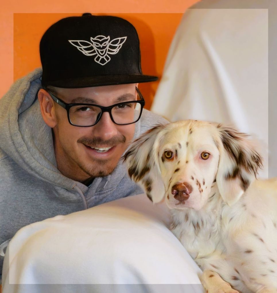 A smiling man in a black owl-embroidered snapback hat and glasses poses closely next to a white and brown spotted Llewellin Setter dog.