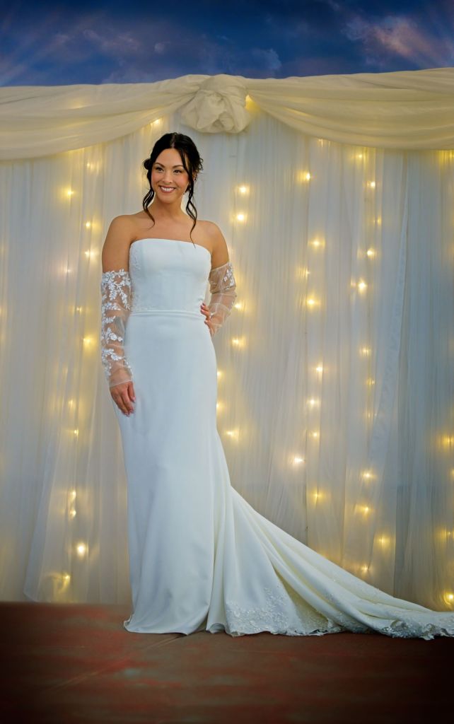 A smiling Jasmine Sonmor models a strapless wedding dress with detached lace sleeves, standing in front of a white tulle backdrop illuminated by fairy lights at the Bigwood Event Center, Fergus Falls MN.