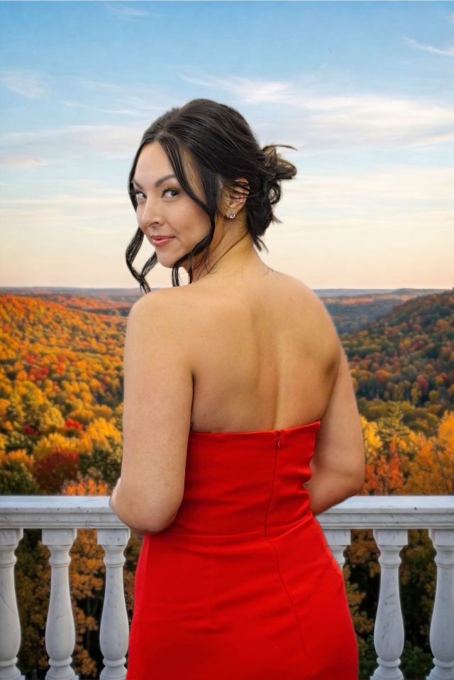 A striking, elegant portrait by Lance Albers. The vibrant pop of the strapless red dress contrasts beautifully with the warm tones of the background, creating a very strong visual anchor. An outdoor portrait of the owner of Aura Fabricators wearing a formal red dress on a balcony overlooking an autumn forest landscape.