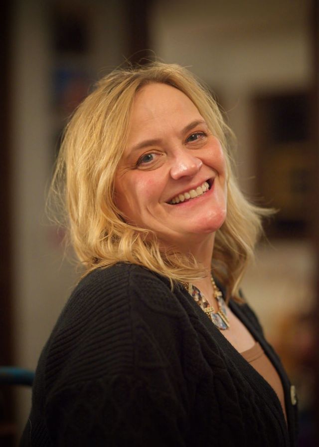 A close-up portrait of local artist Jenny Field smiling brightly, wearing a textured black cardigan and an artistic chunky necklace in a softly lit indoor setting.