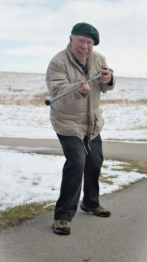 An older man wearing a green beret and a beige winter jacket laughing and playfully pointing a monopod like a telescope outdoors in winter at the Prairie Wetlands Learning Center in Fergus Falls, Minnesota.