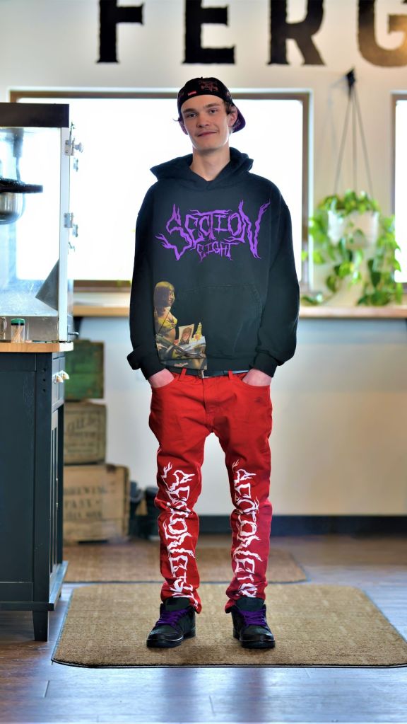 A young high school graduate standing indoors at Fergus Brewing Co wearing a black Section Eight hoodie, red graphic pants, and a backward cap.