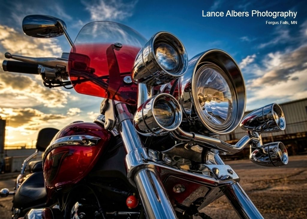 Close-up dynamic front view of a red cruiser motorcycle showcasing polished chrome forks, bright dual headlights, and a translucent red fairing windshield against a cloudy blue sky at sunset.