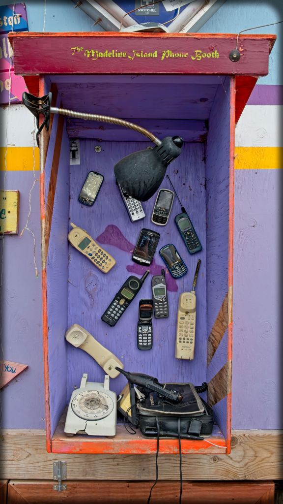 A close-up shot of the open wooden shadow box at The Madeline Island Phone Booth art installation, showcasing a collection of obsolete flip phones, brick cell phones, and a vintage rotary landline telephone.