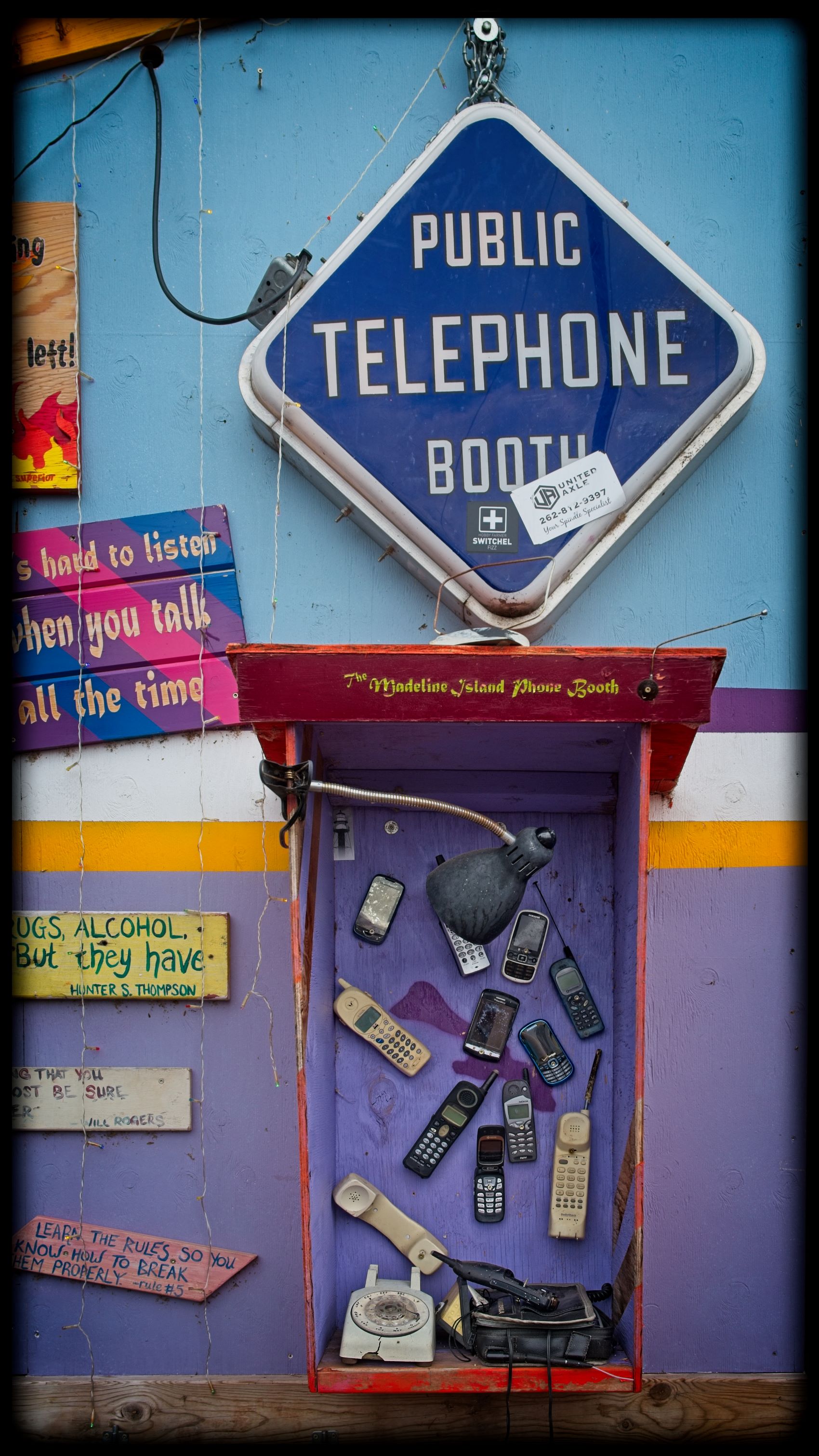 An eclectic outdoor public telephone booth installation at Tom's Burned Down Cafe on Madeline Island, featuring an assortment of vintage cell phones and an old rotary telephone mounted inside a wooden box under a blue sign.