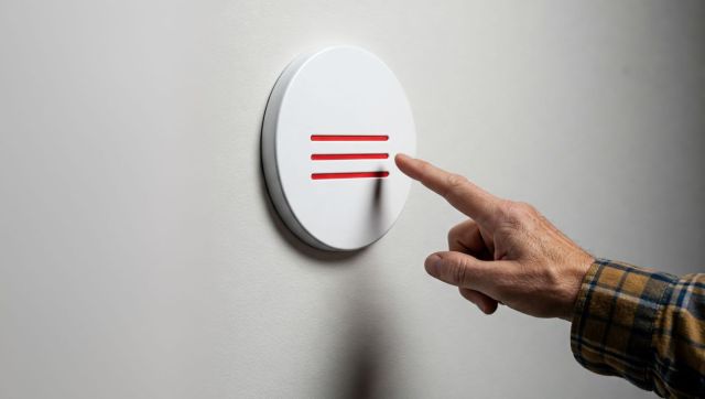 Close-up photo of a hand in a yellow and blue plaid shirt pointing a finger at a white circular button mounted on a wall, featuring three horizontal red stripes resembling a hamburger menu icon.