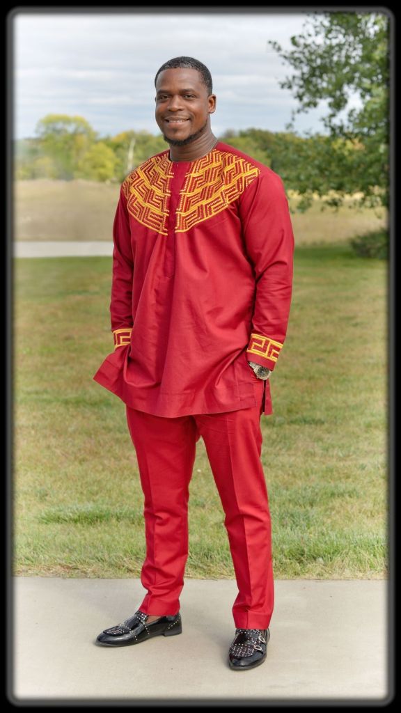 A smiling man wears a vibrant red African dashiki tunic and trousers outfit with yellow geometric embroidery.