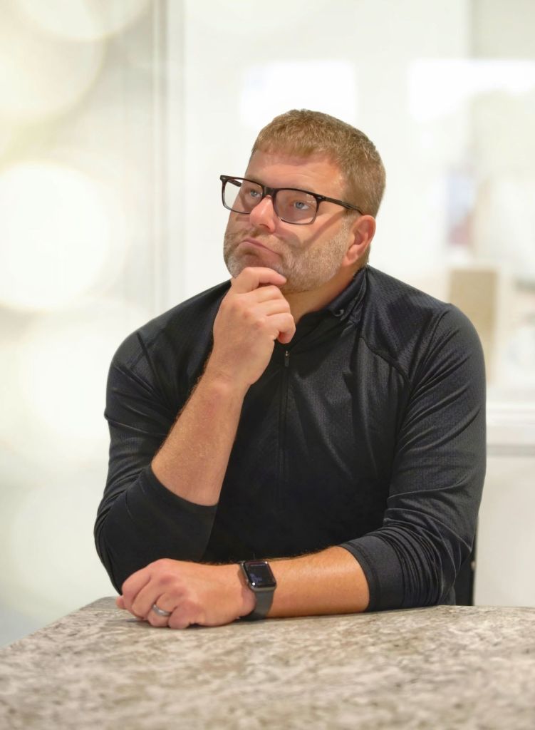 A man with glasses and a short beard sits at a counter, resting his chin on his hand in a thinking pose.