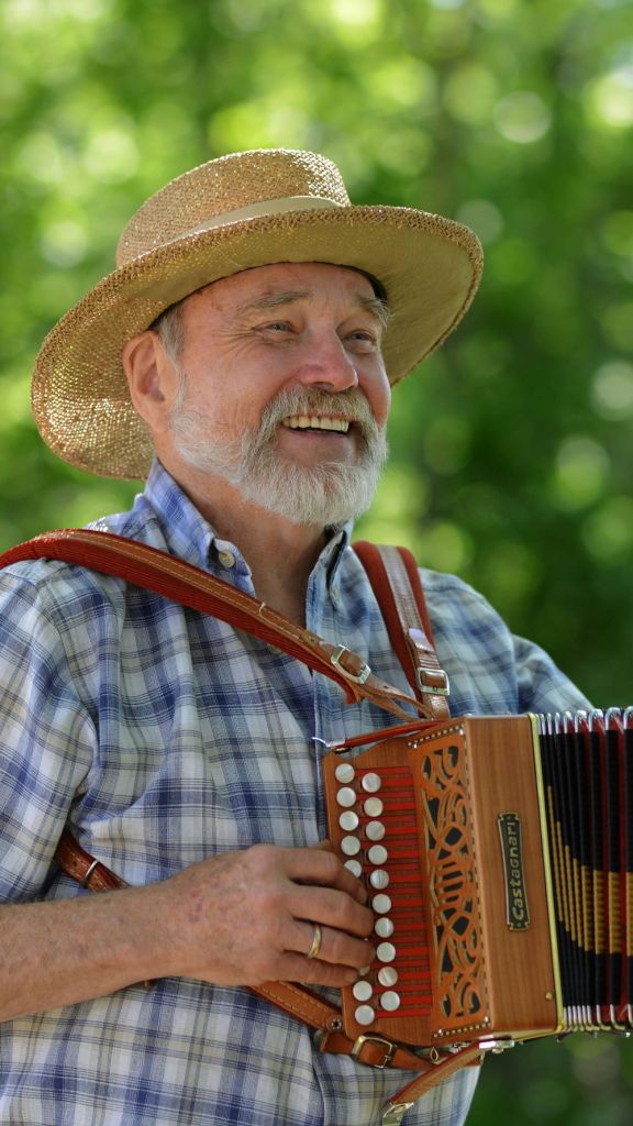 Mikkel Thompson smiling while playing a Castagnari button accordion outdoors, wearing a straw sun hat and a blue plaid shirt against a blurred green background.