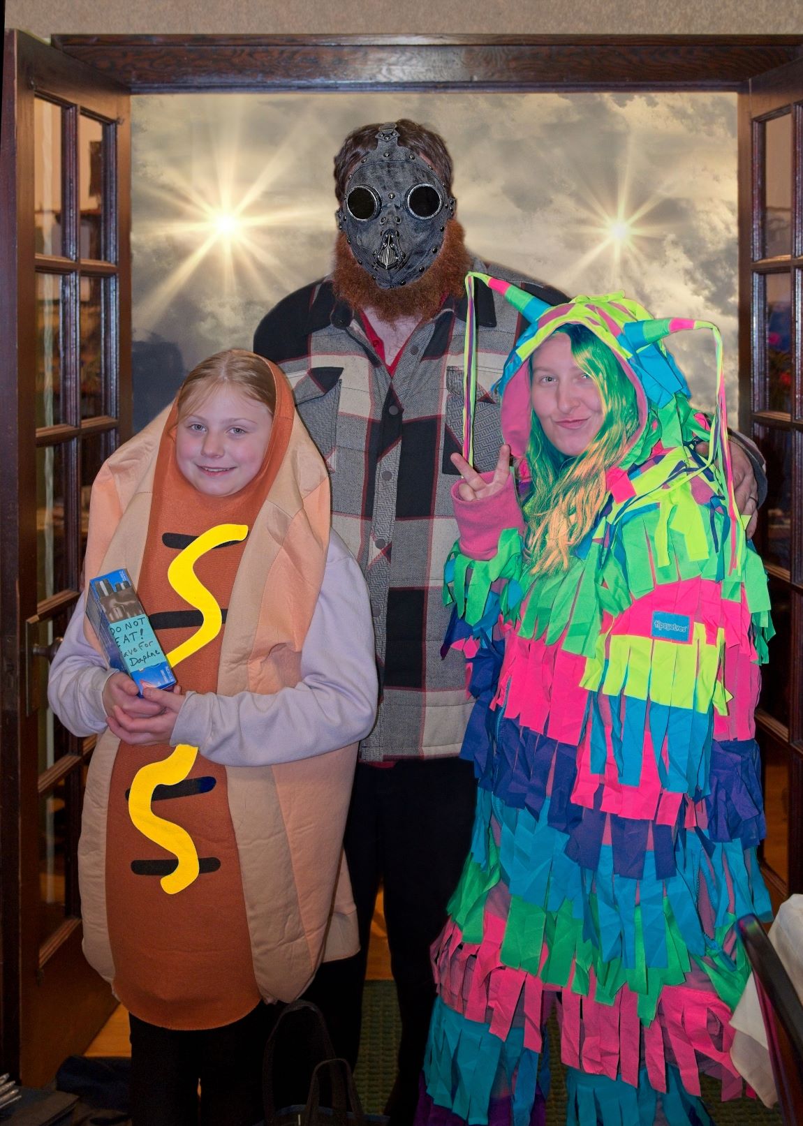Three people posing in Halloween costumes, including a child in a hot dog costume holding a treat box, a woman in a colorful piñata costume, and a man wearing a dark steampunk gas mask and a plaid jacket.