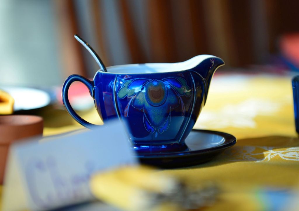 A glossy cobalt blue ceramic creamer pitcher with an ornamental leaf pattern and a silver spoon inside, resting on a matching saucer over a yellow tablecloth.